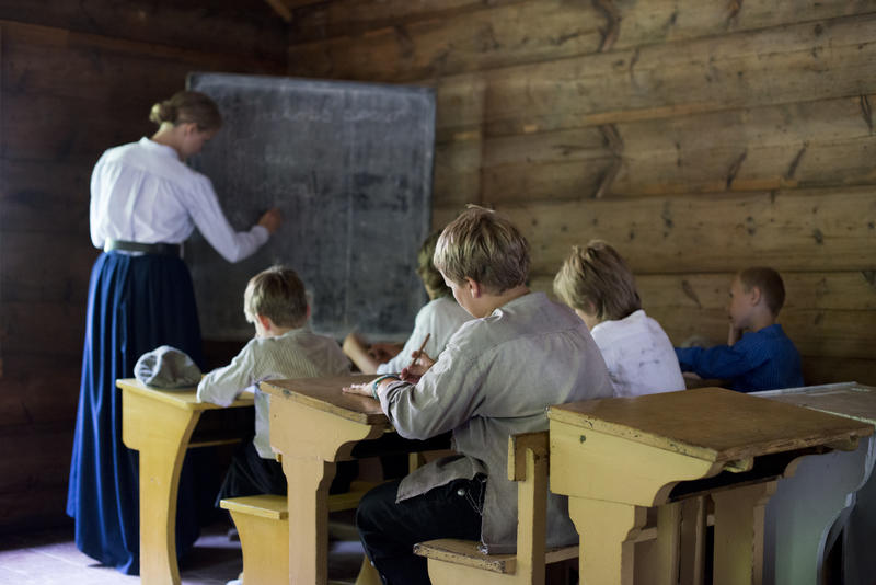 Barn sitter ved pultene i gammel skolstue mens lærerinne skriver på tavlen (Foto/Photo)