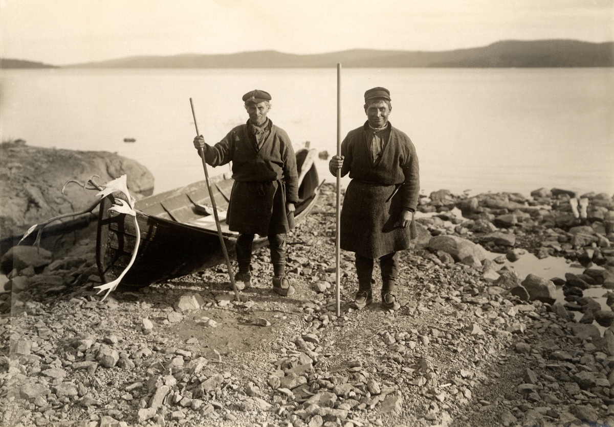Två samer står på stranden vid en sjö, bredvid en eka. I båten ligger ett renhorn.