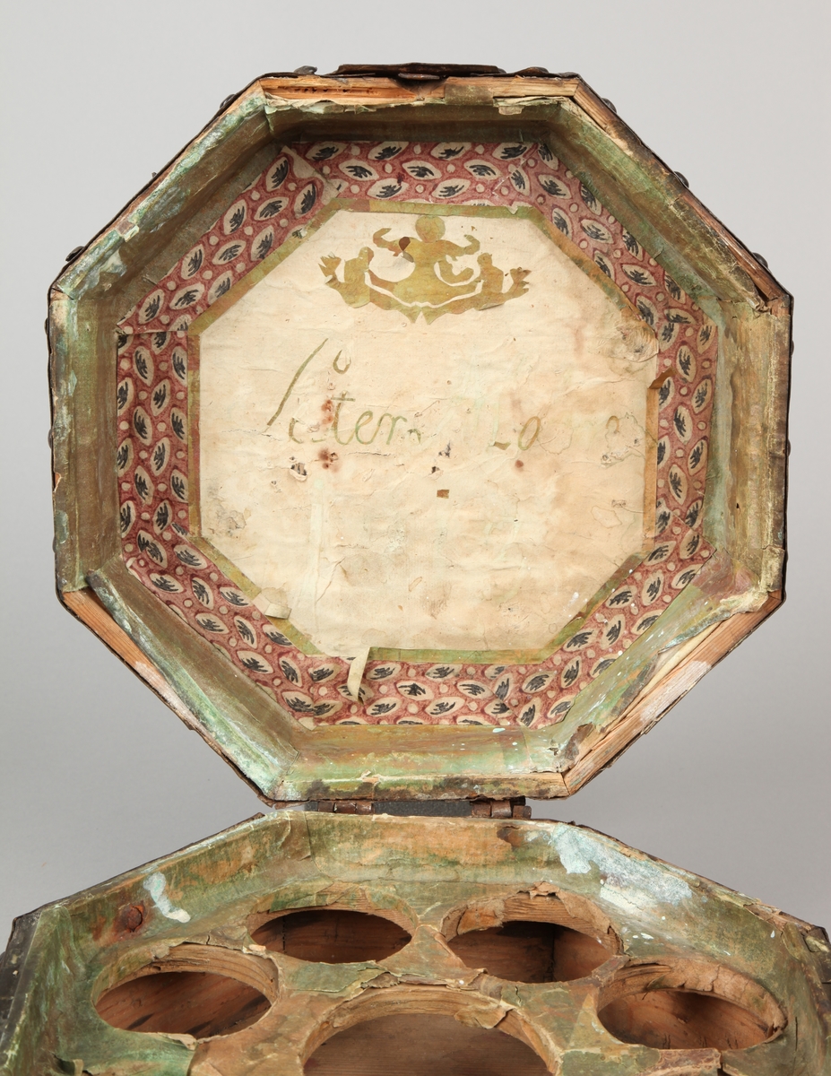 Åttekantet. Den har dekorerte flater i messing med jernbeslag og det hele er naglet på tre. På lokket er det en hank. Flaskefôret er låsbart, men den originale nøkkel mangler.