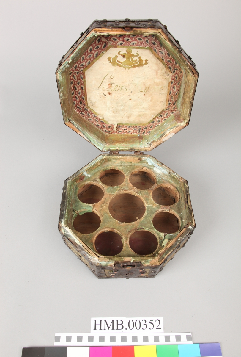 Åttekantet. Den har dekorerte flater i messing med jernbeslag og det hele er naglet på tre. På lokket er det en hank. Flaskefôret er låsbart, men den originale nøkkel mangler.