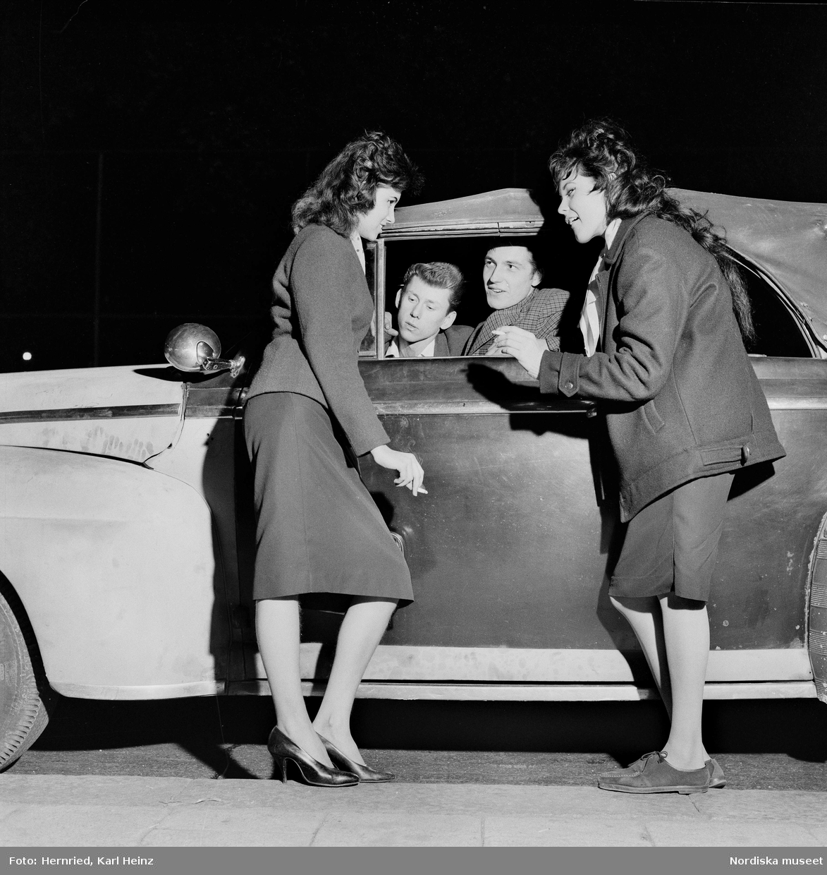 Gatumiljö med bilburen ungdom.Två unga kvinnor, den ena med cigarett i handen, hänger vid bil och samtalar med två unga män som tittar ut genom den nedvevade rutan. 