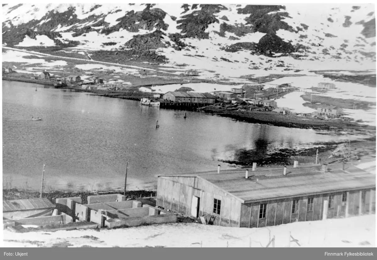 Oversikt over Bergfjord. I forgrunnen kan man se administrativbrakken og omtrent midt på bildet, der båten ligger, ser man en kai og bygning for fiskemottak. Der ligger det også en butikk. Bildet tatt en gang i perioden 1946-48. På bildet kan man se hus med vinduer og piper. I bakgrunnen ser man fjell og snø.