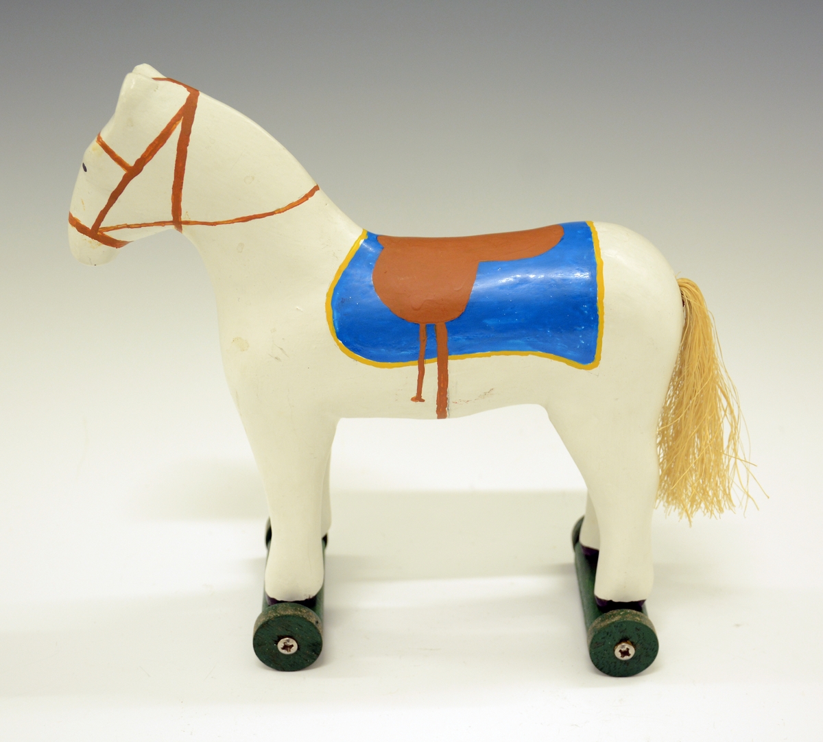 Trehest på hjul. Hesten er malt hvit med blått dekken, og brunt seletøy. Hesten har hale av hamp.Under beina er det skrudd på 4 stk grønne hjul.