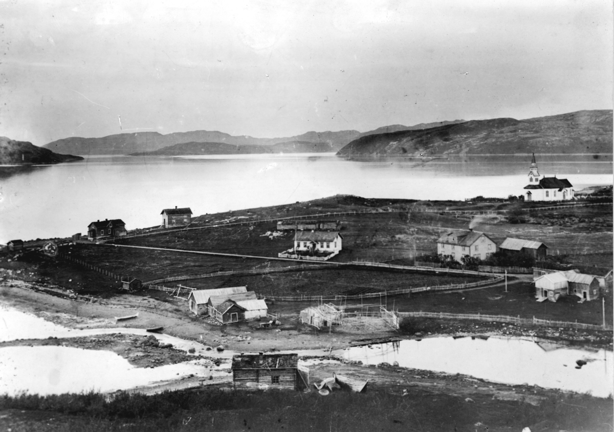 Oversiktsbilde fra Kirkenes i 1902. Båter ved stranden, noen fiskehjell og naust. Tre/ fire gård og kirken til høyre.  Bildet er en reprokopi (en bra en) laget av M. Karlsen i 1952.