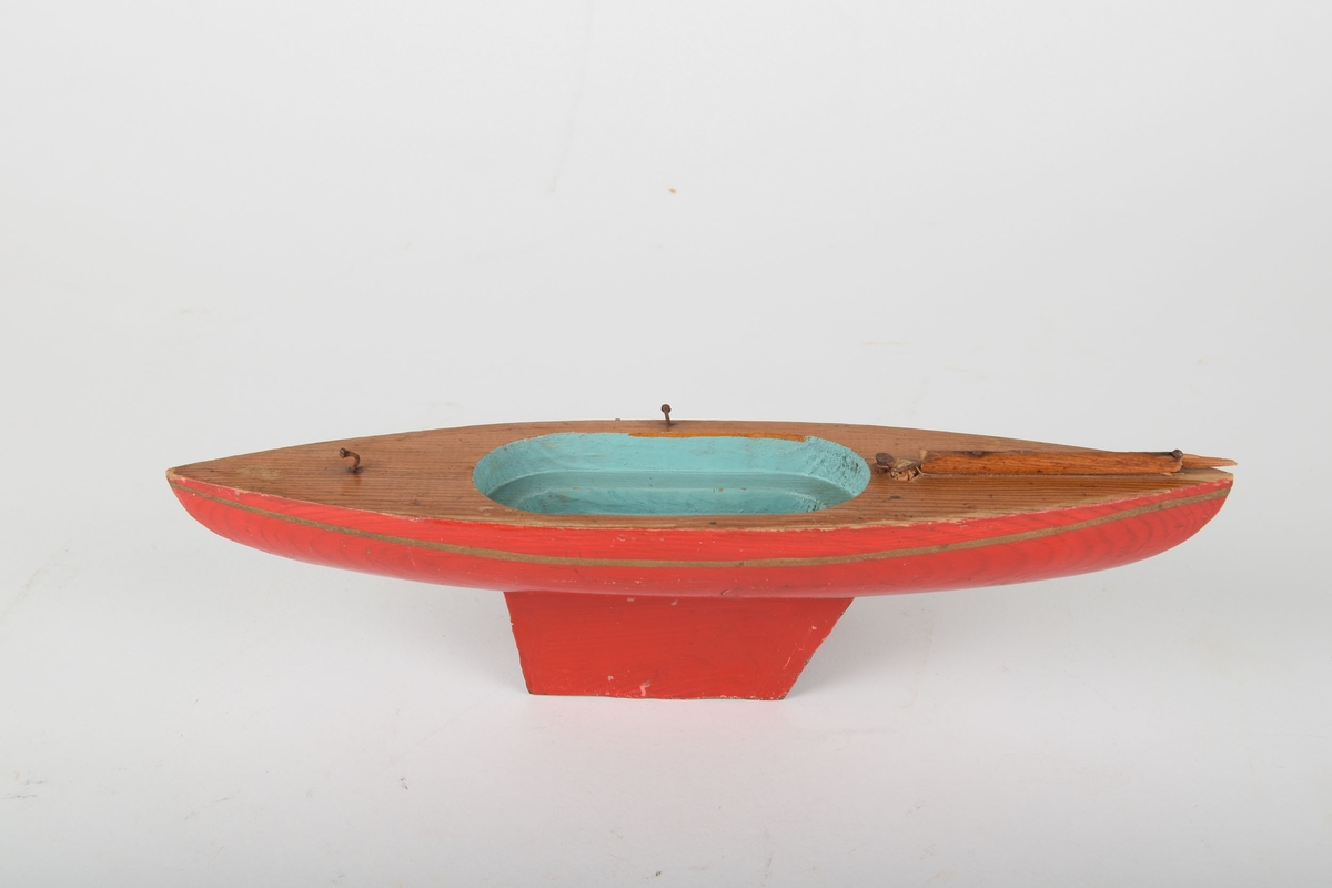 Lekebåt i tre. Rød utvendig, med trefarget dekk. Innsiden er turkis. Båten har 3 metallfester og 1 pinne på dekket.