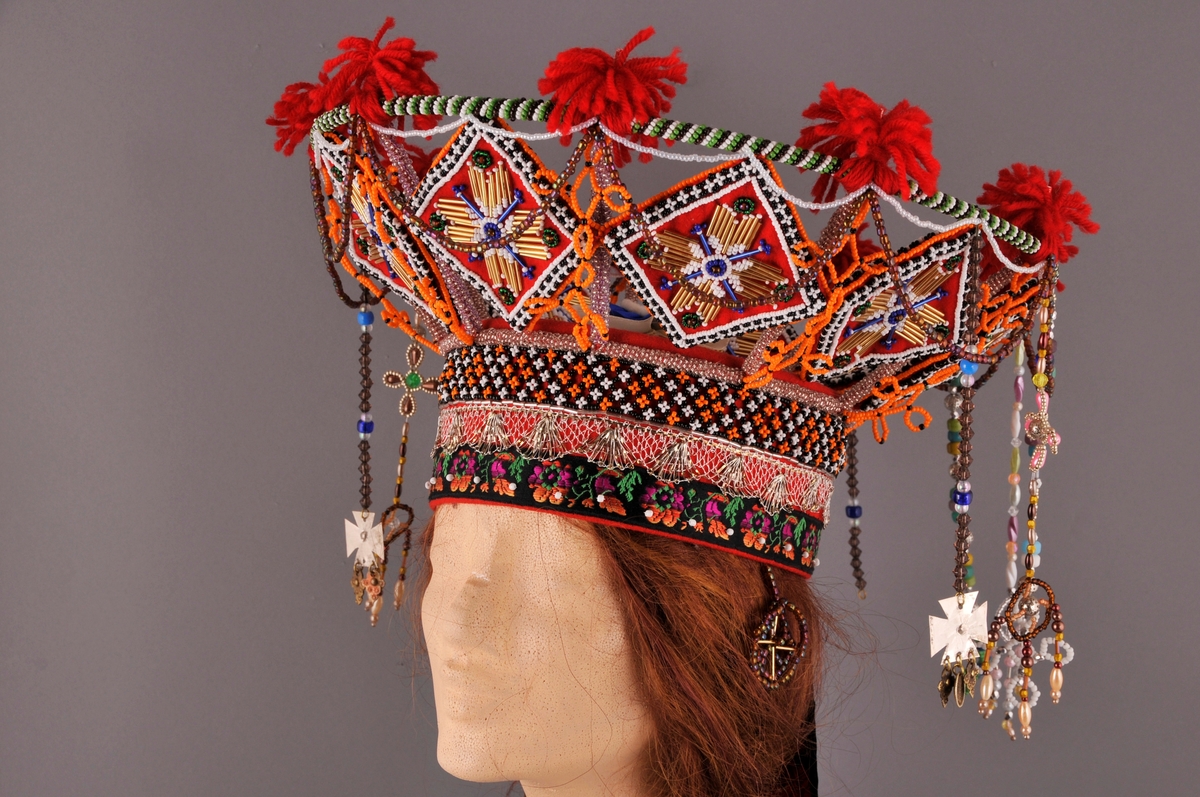 Brudekrone, nylaga.
"Pull" truleg med botn av stiv kartong og kledd med kraftig raudt klede. Påsydd (frå nedkant og oppover) eit band med satengbotn lansert blondemønster og påsydd kvite glasperler, ein sylvknipling og eit perlebendel. Perlebendelet er tredd med glasperler i svart, kvitt og orange.Sjølve krona er oppbygd rundt eit stativ av stål, raudlakkert. Heile stativet er dekt av glasperler i ulike fargar. Inni kvart "rom" er det eit sydd raudt kvadratisk klede som er pryda med glas- og stavperler av åttebladrose-mønster på begge sider, alle kvadrata er like og dei er montert på høgkant. I tillegg er det ein del ulike heng frå øvre kant av krona, her er det nytta ulike typer glasperler. På toppen av krona er det festa små duskar av laustvinna, raudt ullgarn i kvart skjæringspunkt. Inni krona, over hovudet, ligg eit ubleika band i kryss og i krysset er det sydd fast ein rosett.
Bak på krona er det sju heng av ulike band. På kvar side er det "øyrepynt". Frå sida og bakover er det 11 heng med ulike typer perler.