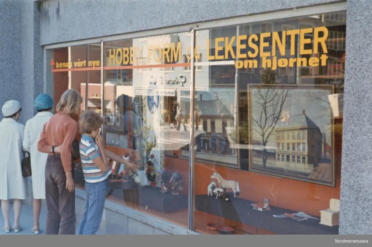 Foto fra Sverdrups bokhandel i jubileumsåret 1974, da bedriften feiret sitt 100 års jubileum. Her ser vi to gutter og to voksne damer som kikker inn på et av utstillingsvinduene ved butikken. Fra Nordmøre museums fotosamlinger.

