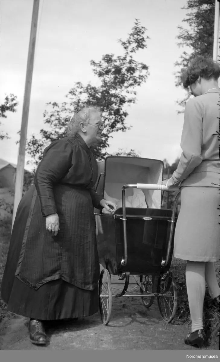 Foto av Margrethe Olise Sverdrup (født Barman)  stående til venstre i bildet. Trolig er det et av Margrethe Sverdrups barnebarn vi ser i vognen. Datering er trolig omkring 1920 til 1939. Det er usikkert hvor bildet er fotografert, men kan være fra Eidsvåg i Nesset kommune. Fra Nordmøre museums fotosamlinger. EFR2015