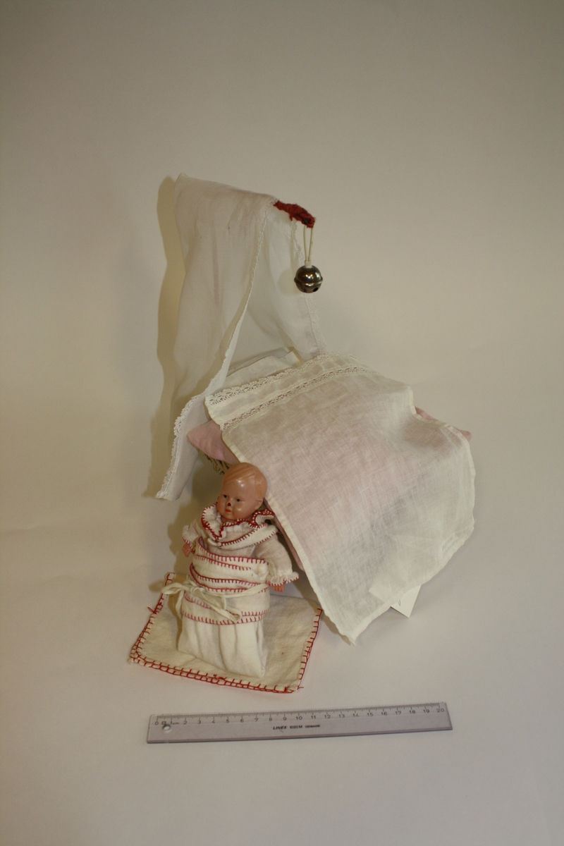 Celloloid-dukke i kurv med hjemmesydd sengetøy og sengeforheng og hjemmelaget stativ med bjelle. Dukken er reivet har har tøybleie og skjorte med krage.