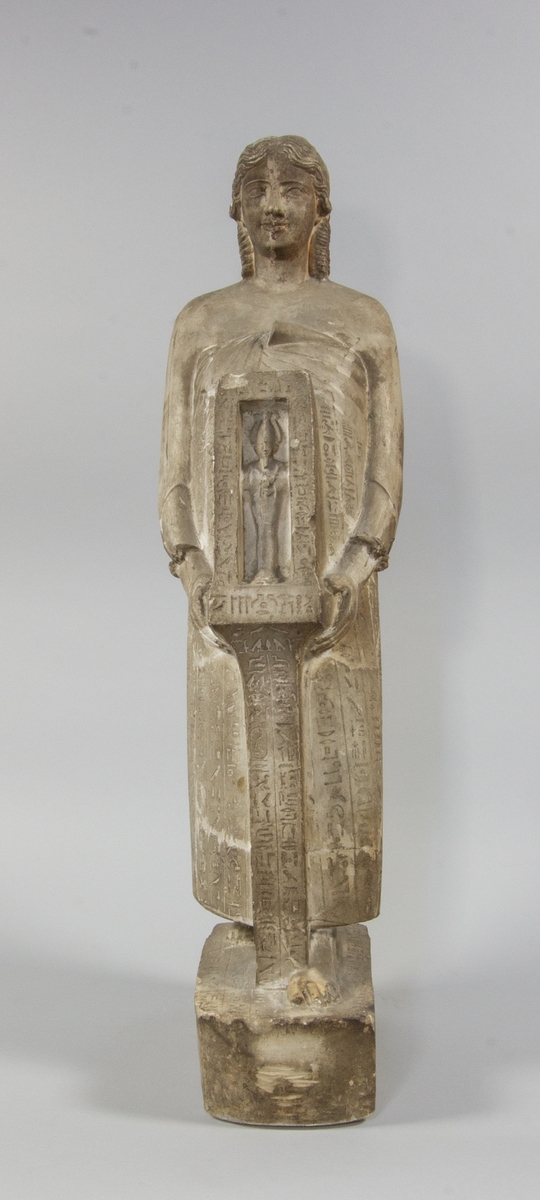 Kvinnogestalt stående i helfigur i en egyptisk pose med fötterna i en rak linje. Klädd i lång klädnad med bara axlar. Hon håller med båda händerna en stående rektagulärt öppen sarkofag innehållande en möjlig mumie av en farao. Ytorna täckta med hieroglyfer.