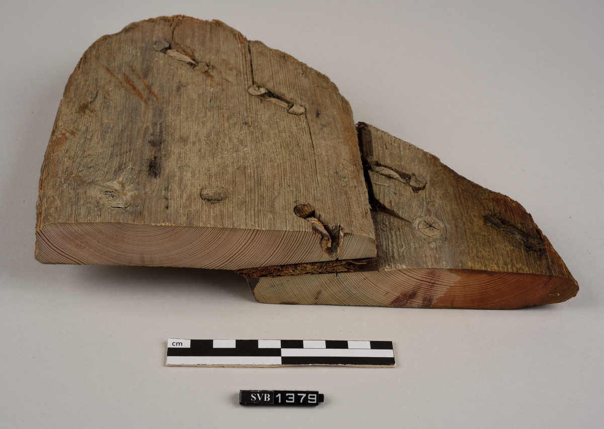 Del av et båtbord. Delen ble saget av. Det er hull til vidje og plugger. Delvis er det rester av både vidje og plugger. Treet fremstår ubehandlet. Det er et materiale som ligner torv/mose mellom bordene.