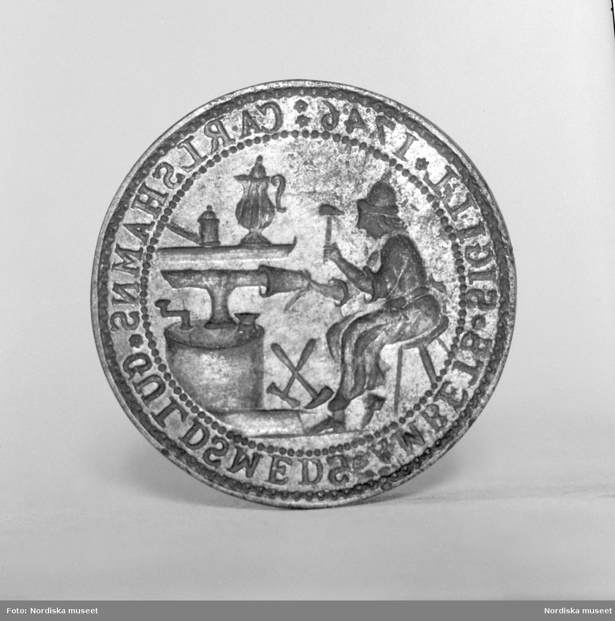 Sigillstamp av järn från 1746 som tillhört Karlshamns guldsmedsämbete. På stampen graverat en man sittande vid ett städ hamrande på en bägare. Runt kanten texten CARLSHAMNS GULDSMEDS ÄMBETS SIGILL 1746.