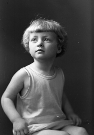 Foto av en okänd liten flicka i ett enkelt linne.
Knäbild. Ateljéfoto.