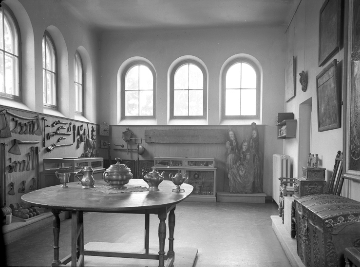 Interiör från Smålands museum. Avdelningen för allmoge-högrestånd.
Till höger syns bl.a en kista, en karmstol och på väggen hänger ett antal tavlor. I mitten syns bl.a. nåt slags skål och en kanna/stop av tenn på bordet.
Till vänster hänger bl.a. ett antal yxor, något slags pistoler.
