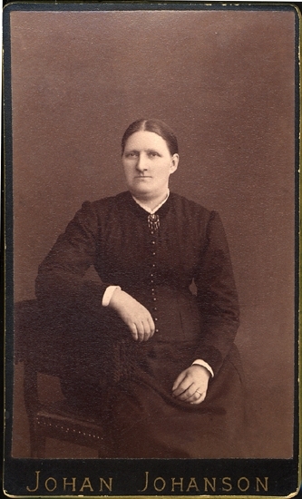 Porträtt (knäbild, halvprofil) av en okänd sittande kvinna i mörk klänning med en brosch med kläppar i halsgropen.
Hon sitter på en stoppad stol, prydd med tännlikor och korta fransar.