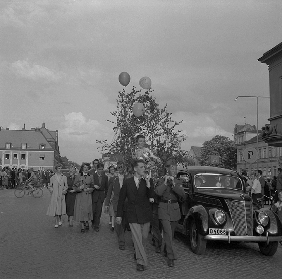 Studenterna, tredje dagen, 1953. 
En kvinnlig student bärs hem i lövad bärstol från Stortorget. I bakgrunden syns några av husen längs Kronobergsgatan.