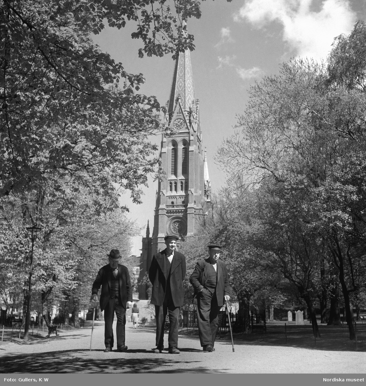 Norrmalm, Stockholm. Tre äldre män tar en promenad i solskenet. Johannes kyrka i bakgrunden.