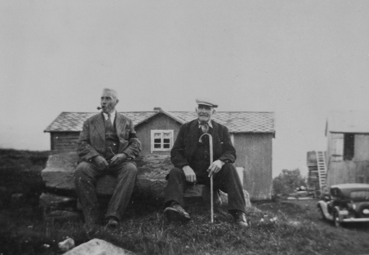 To eldre karer sitter på en stor stein foran bygningene på gammelgården på Klasvollen ved Aursunden