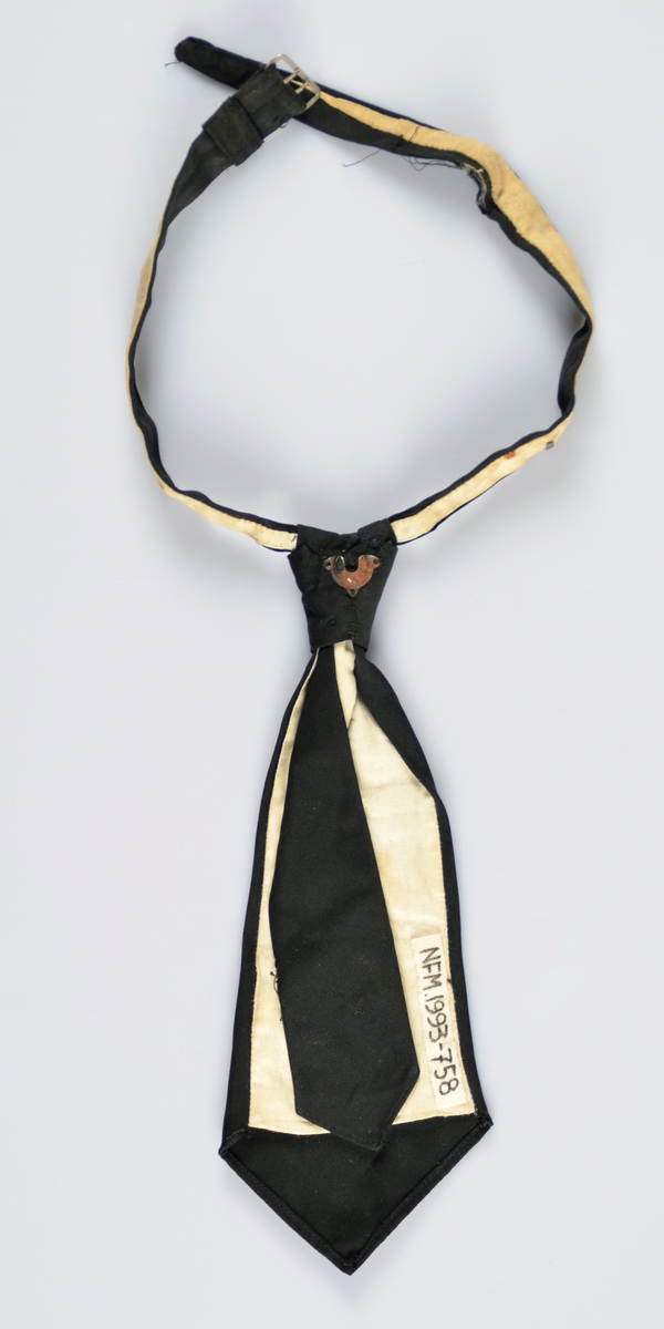 Kort, ferdigknytta slips. Svart med kvitt fôr. Truleg silke. Lukking i nakken med metallspenne. Metall ting bakside knute, 2 metallhemper halsband.