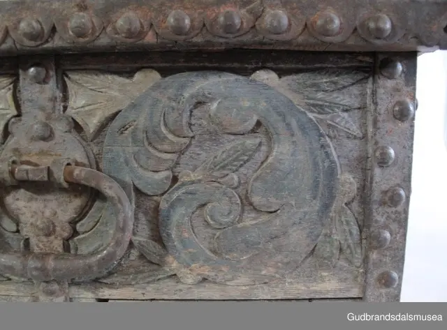 "Bladformer, smøygde band og drageornamentikk. Millom kantjarn og utskurden hev lokket fint utskorne små blad og halvrunde aggar. Vel vedlikehalde, sterk jarning og prydelege gangjarn. Truleg frå 1500 talet. Fra Haugen, Øystre Gausdal". (Jf. "Katalog for Lom Bygdesamling,1925).
Skrinrammen er holdt sammen av utvendige hjørnekantbeslag; jernhandtak i hver side. Fronten har nøkkelhullslås med beslag rundt. Lokket er satt samen av 2 bord, forsterket med jernbeslag rundt kanter på overside; to hengselbeslag festet til innside av lokk og bakstykke. Skrinsider blir holdt sammen av beslag ned langs hjørnekanter; har jernhandtak festet på jernbeslag.
Bunnen er satt sammen av 2 bord, festet til hverandre med to forsenka treplugger; bunnen var opprinnelig festet til underkant av skrinsider med 4 x 4 trenagler,seinere festet med spiker. 
Det er jernbeslag  på bakstykket midt, rundt hjørnekanter, omkring nøkkelhullslås, rundt lokk samt 2 handtak med beslag. Alle beslag er festet med store jernnagler; beslag på sidestykker er festet med små jernnagler under skrinbunnen. 3 av 8 bunnfester for beslag er fortsatt intakte.
DEKOR. De store jernnaglene med forskjøvet plassering på beslag ble sannsynligvis brukt som dekorelement; runde beslag på sider, bakstykke og front. Flatskurd på lokk med flettet, stilisert renessanseranke, der bladverk er "omgjort" til småspiraler eller rundinger.  På skrinsidene akantusskurd med c-former, bladtunger uten stilk og naturalistiske småblad i rokokkostil. Flatskurd var vanlig i Gudbrandsdalen i perioden ca. 1650-1750, mens akantusmotivet først ble vanlig på bruksgjenstander i løpet av siste halvdel av 1700-tallet. Den mørkere grønne og blå dekorfargen var vanlig i siste del på 1700-tallet; den brune bunnfargen var vanlig i bruk fra 1780-tallet. 
BRUK. Før kofferten kom i bruk hadde man reiseskrinet, som ble brukt til oppbevaring av bl.a. klær, verdisaker og mat på reiser. Skrinet hadde ofte et eller to mindre, smale rom langs den ene skrinsiden og ett større rom. De fleste reiseskrin var i tillegg forsterket med jernbeslag langs alle kanter og med en solid nøkkelhullås til sikring mot støt, slag og tyveri.