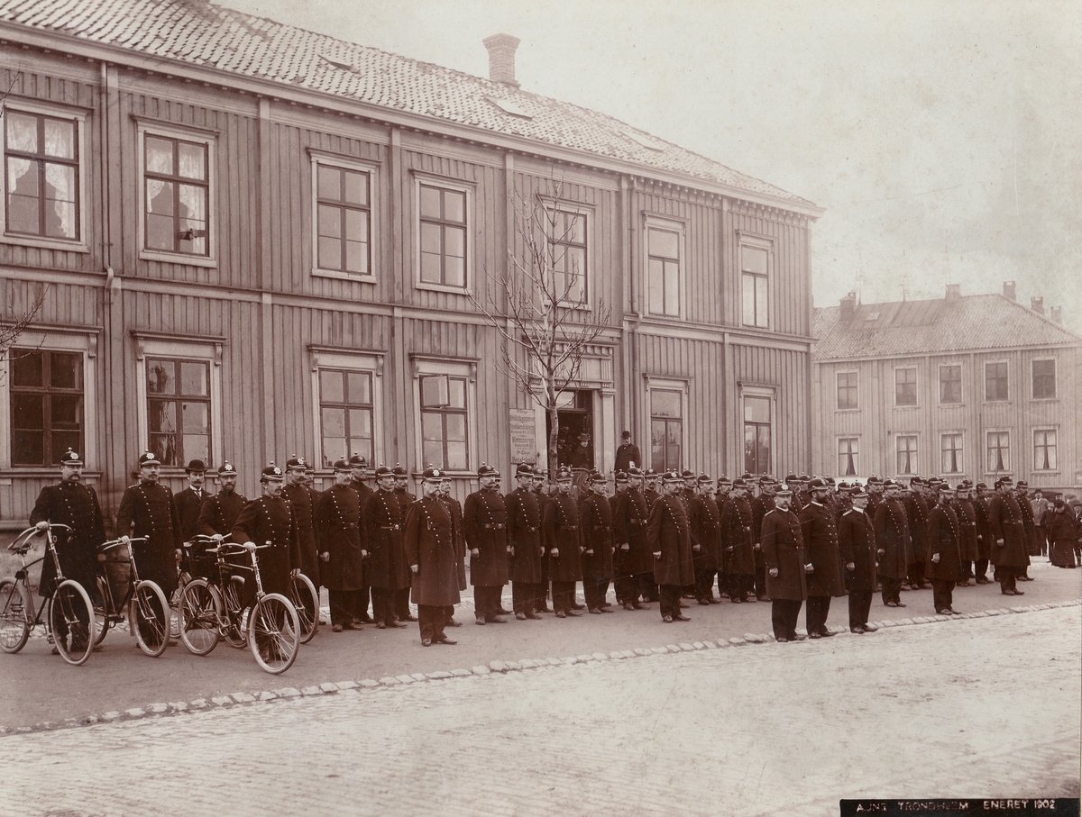 Trondheims politistyrke oppstilt foran politikammeret i Hornemannsgården på torvet i Trondheim. Alle er iført uniformsmodell 1895, og noen har sykkel.