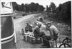 Tre mann sitter på melkespann i veikanten. Buss i venstre bi