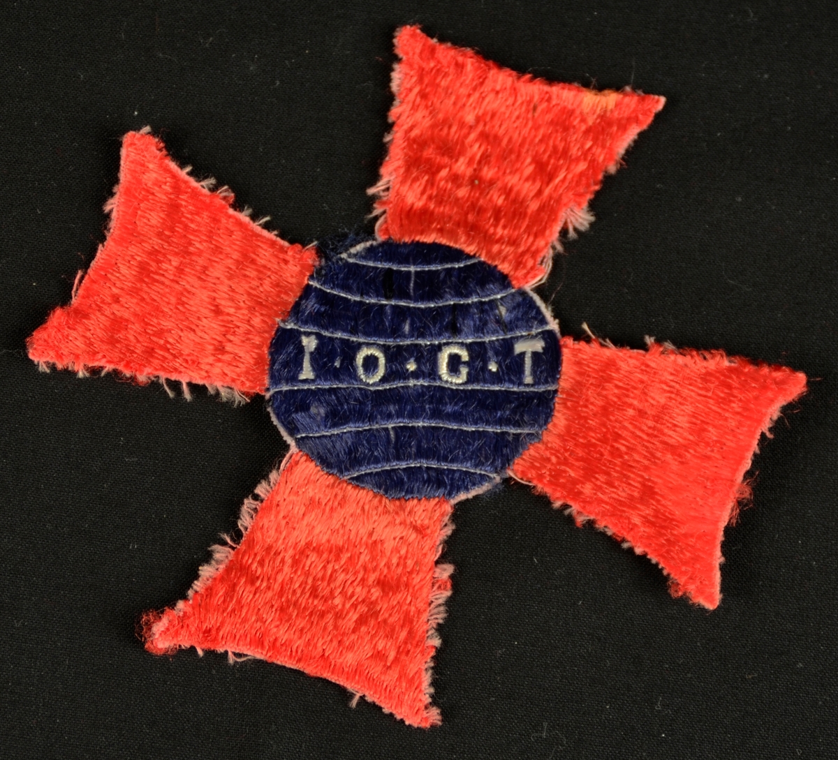I.O.G.T. losjemerke i tekstil. Kross-form. Blå sirkel med kvite bokstavar og stripar, 4 raude vingar.