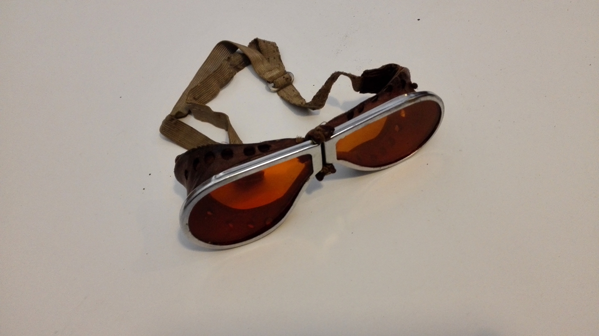 Heildekkande solbriller med oransje glas, med metallramme og skinn rundt, slik at brillene sit tett mot huda kring heile auget.
Brillene vart festa med regulærbar strikk bak nakken.