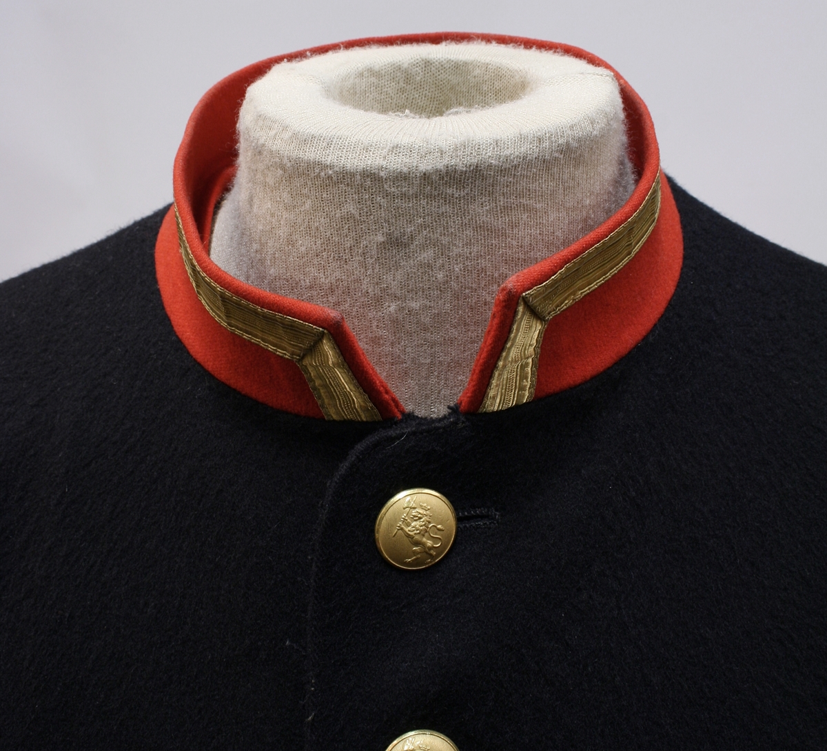 Rød krage med gullkant, riksløveknapper (enkeltknepper), splitt bak med seks knapper og rød paspoil.