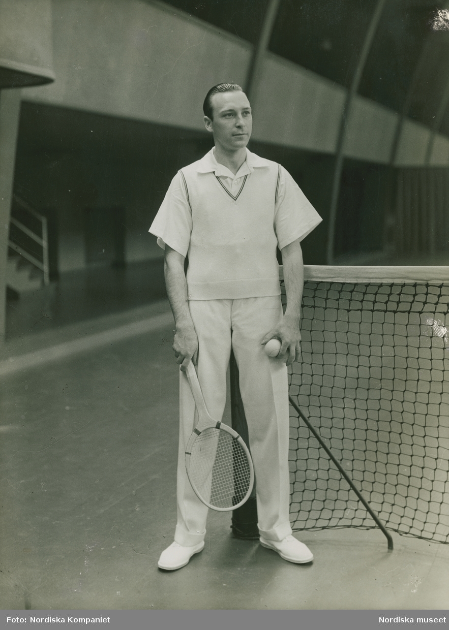 Man i vit skjorta, vit slipover och vita byxor bredvid ett nät. I ena handen ett tennisracket, i den andra en boll. Nordiska Kompaniet. Text med blyerts på baksidan: "M-sekt. div. bilder.sept 1930"