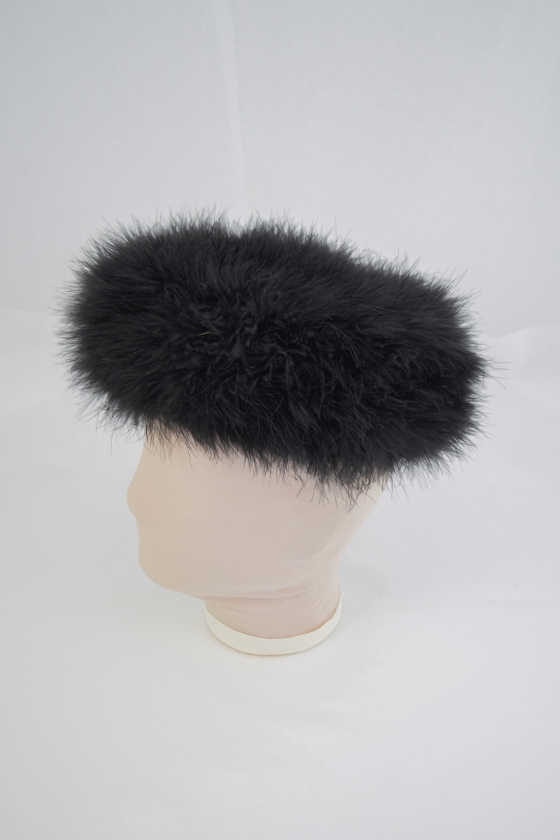 Rund ensfarget hatt med utside av lange tynnne dun. Foret med mørkt og blankt stoff.  Form: sirkulær m/ kant.