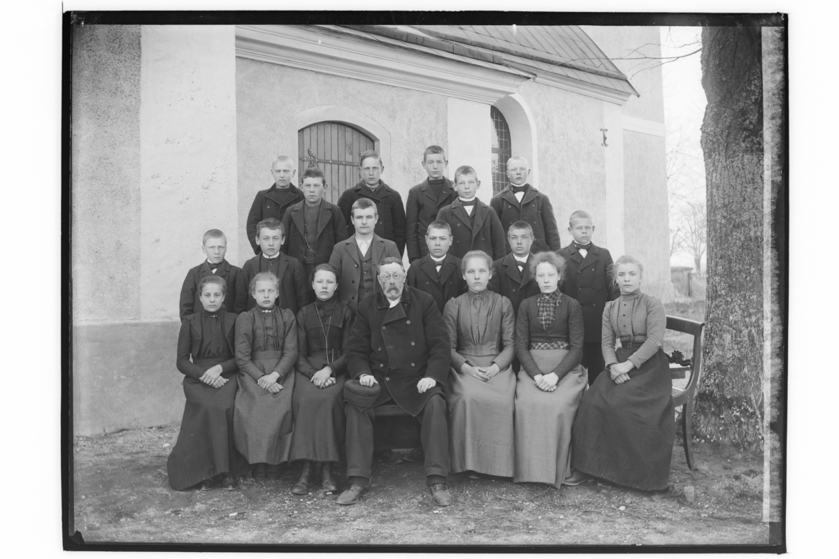 Rinkaby kyrka i bakgrunden.
Konfirmander, 18 ungdomar och en präst.