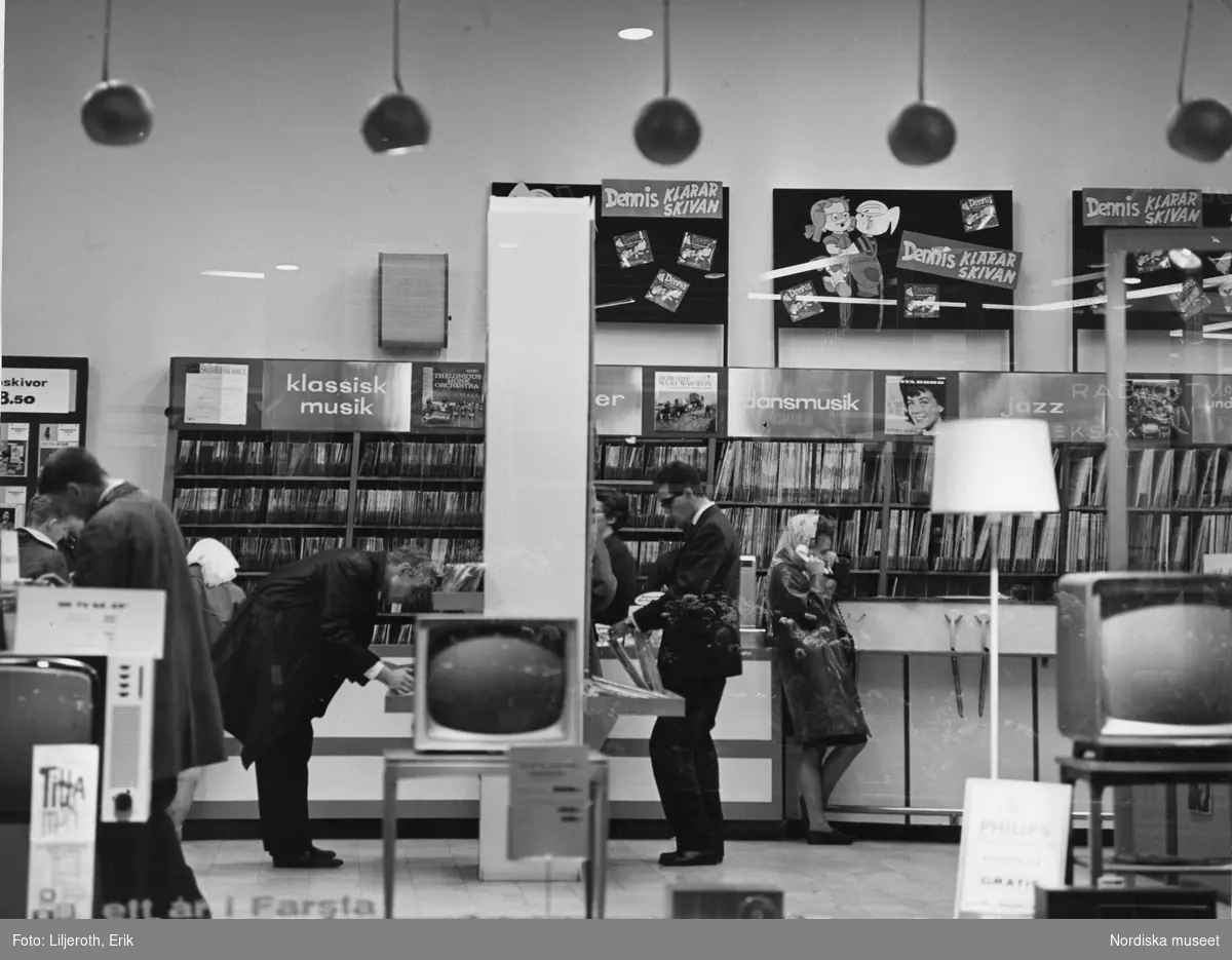 ”Farsta”. Kunder letar grammofonskivor i Radio- & TV-butik. Vid hyllan invid väggen står en kvinna och lyssnar i de lurar som finns  för att provlyssna på en skiva innan köp.