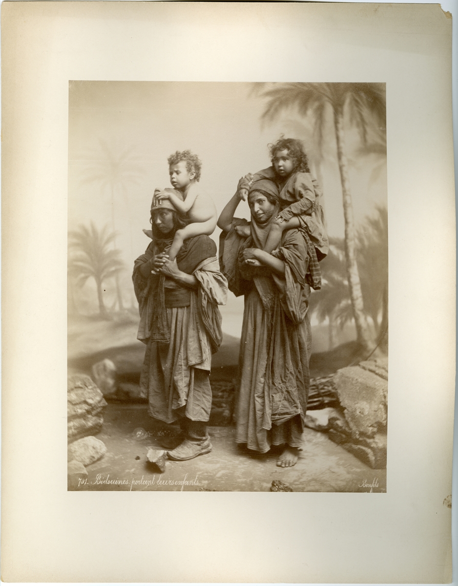 Porträtt av två kvinnor bärande två små barn. Ateljebild med bakgrund, kvinnorna är eller ska föreställa beduiner.