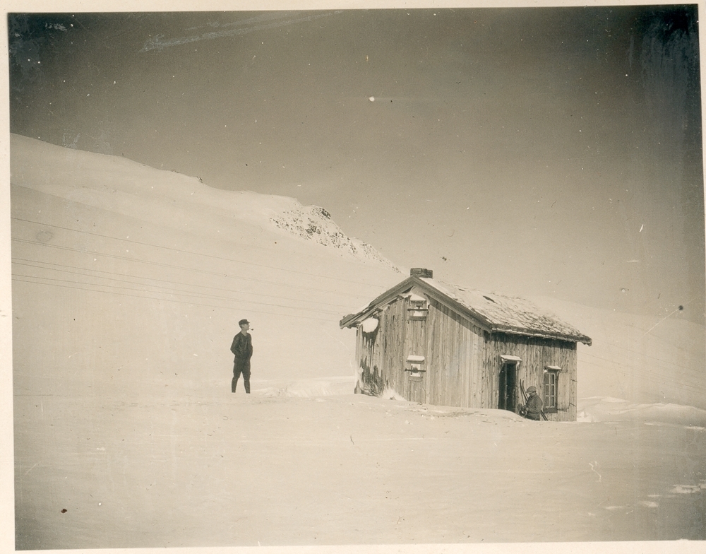 To menn står ved en liten fjellhytte eller fjellstue. Telegrafstue. Snø, høyfjell.