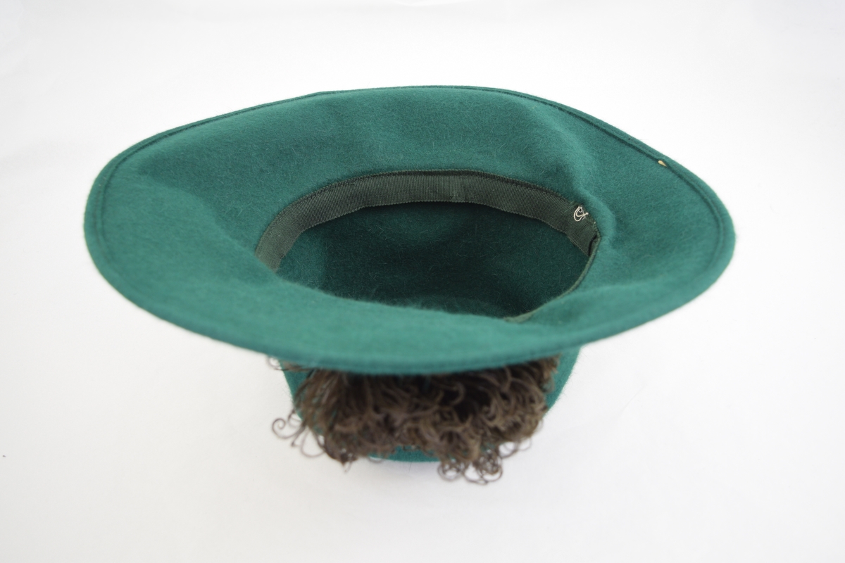 Filt, grønn, maskinsøm. Bred brem krummet opp på en side, pull sylinderformet. I overgangen mellom pull og brem er lagt et grønt bånd av samme materiale. Båndet lagt i en løkke bak og de to endestykkene er festet løst på bremmen. På det samme bånd er festet en "rosett" av svarte strutsefjær, hattens front. På innsiden av hatten er sydd et mørkere grønt hattebånd i hodeåpningen.