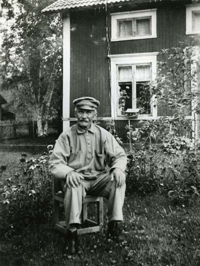 En skomakare i Glösbo, avbildad i sin trädgård. Det är sommar och han sitter på en låg stol framför huset.