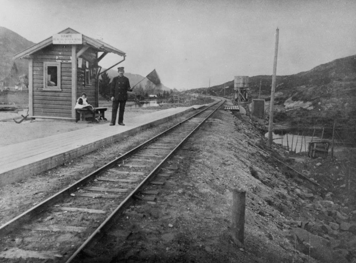 Hamre stasjon på Nesttun-Osbanen. Banevaktar Olaf Fredriksen med flagg. Dottera hans, Magda (f. 1896), sit på benken.
