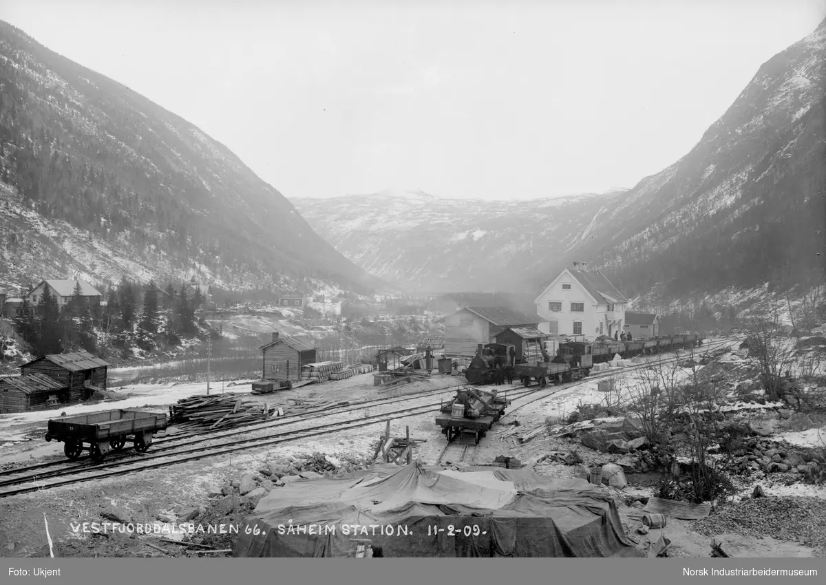 Anleggsområde ved Såheim stasjon. Pakkhus, Såheim stasjon (Rjukan stasjon) og lagerbygg ferdig bygget. To tvilling våtdamplokomotiver stående med vogner på jernbaneskinnene. Tønner liggende foran bygg vest for stasjonen. Mennesker stående foran stasjonsbygning og ved lokomotiver. Noen boliger rundt Rjukan er bygget.