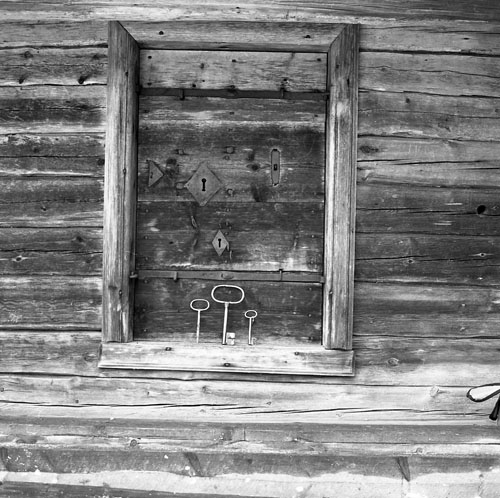 Utsidan av en dörr med ett flertal nyckelhål och tillhörande nycklar som står uppradade på dörrtröskeln nedanför. Dörren fotograferades i Fågelsjö 20-21 oktober 1982.