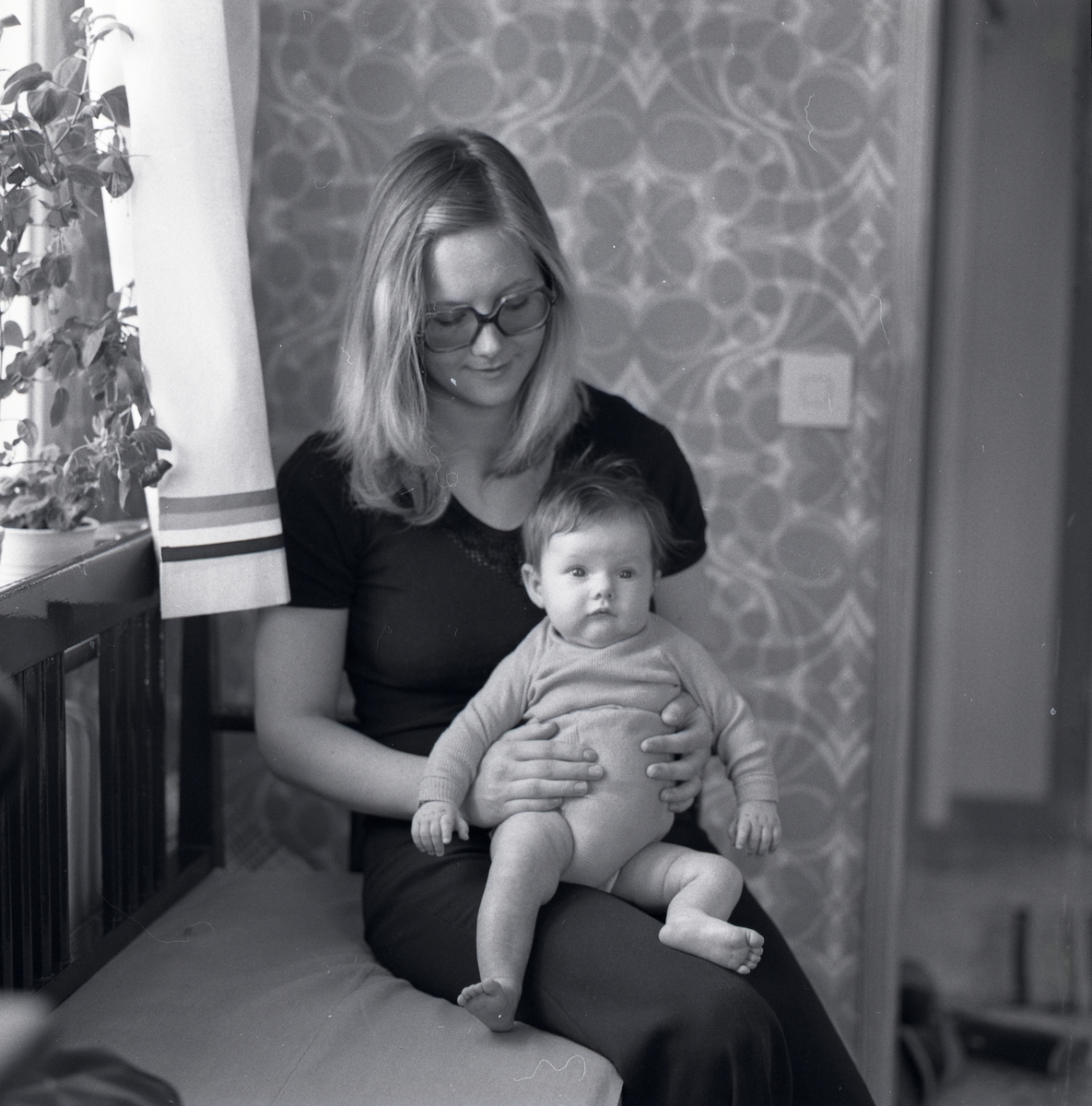En kvinna sitter med en bebis i famnen i ett hus i Flästa, 1975.