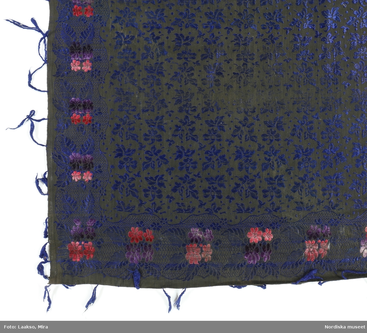 Kvadratiskt halskläde i halvsiden, botten svart bomullssatin, invävt mönster i silke med långa flotteringar. Mittspegl med mörkblå strödda blomkvistar och prickar, kantbård med parställda små enkla blommor i rosa och lila broscherade mot en spetsbotten i blått, klädet blir enkelsidigt och inte vändbart. . Invikta och tråcklade traskanter, 2 stadkanter, Mycket glest iknuten frans av mörkblått silke 
Anm. Tyget skörat vid ena kanten. Sannolikt inte svensk tillverkning.
/Berit Eldvik 2011-12-09