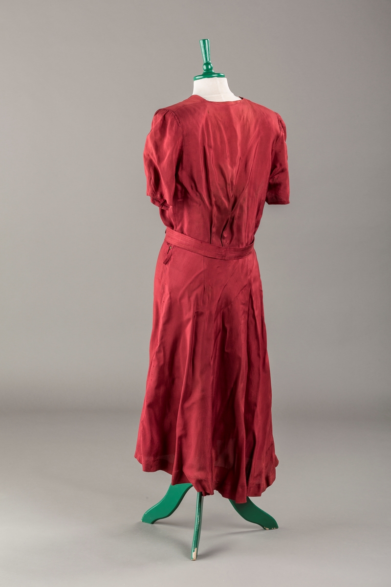 Form: Glatt, klokkeformet skjørt. Forstykkene rynket til buede bærestykker foran. Overdel rynket til skjørt. Korte ermer. Lukket med trykknapper. Belte fastsydd.