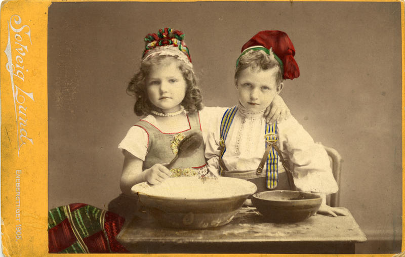 Kolorert studiofotografi av jente og gutt med drakt, sittende ved bord med to treboller, ei sleiv. 1905 (Foto/Photo)