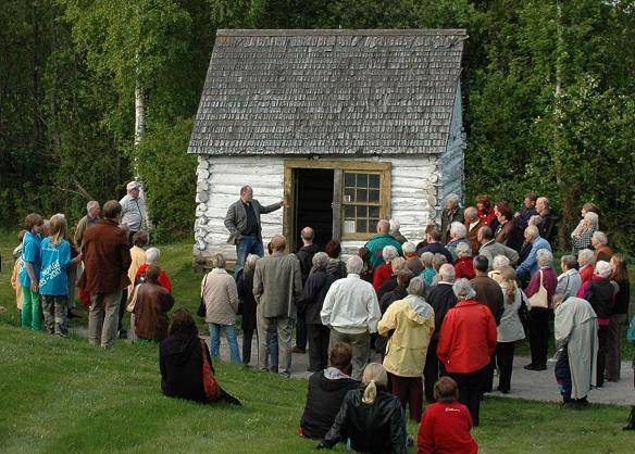 En mann står utenfor et lite, hvitt tømmerhus og forteller om Gundersenstua. Det står en stor gruppe mennesker rundt han.