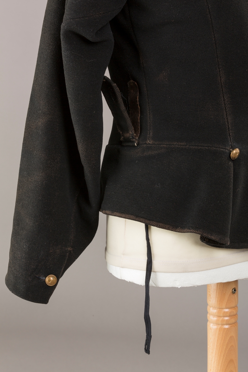 Form: Enkeltspent.

Foret med lin. Forsterket med silke på et lite stykke på ryggen. 12 messingknapper med mønster og tallet 1. Hempe i lin. Bendelbånd med knappehull.