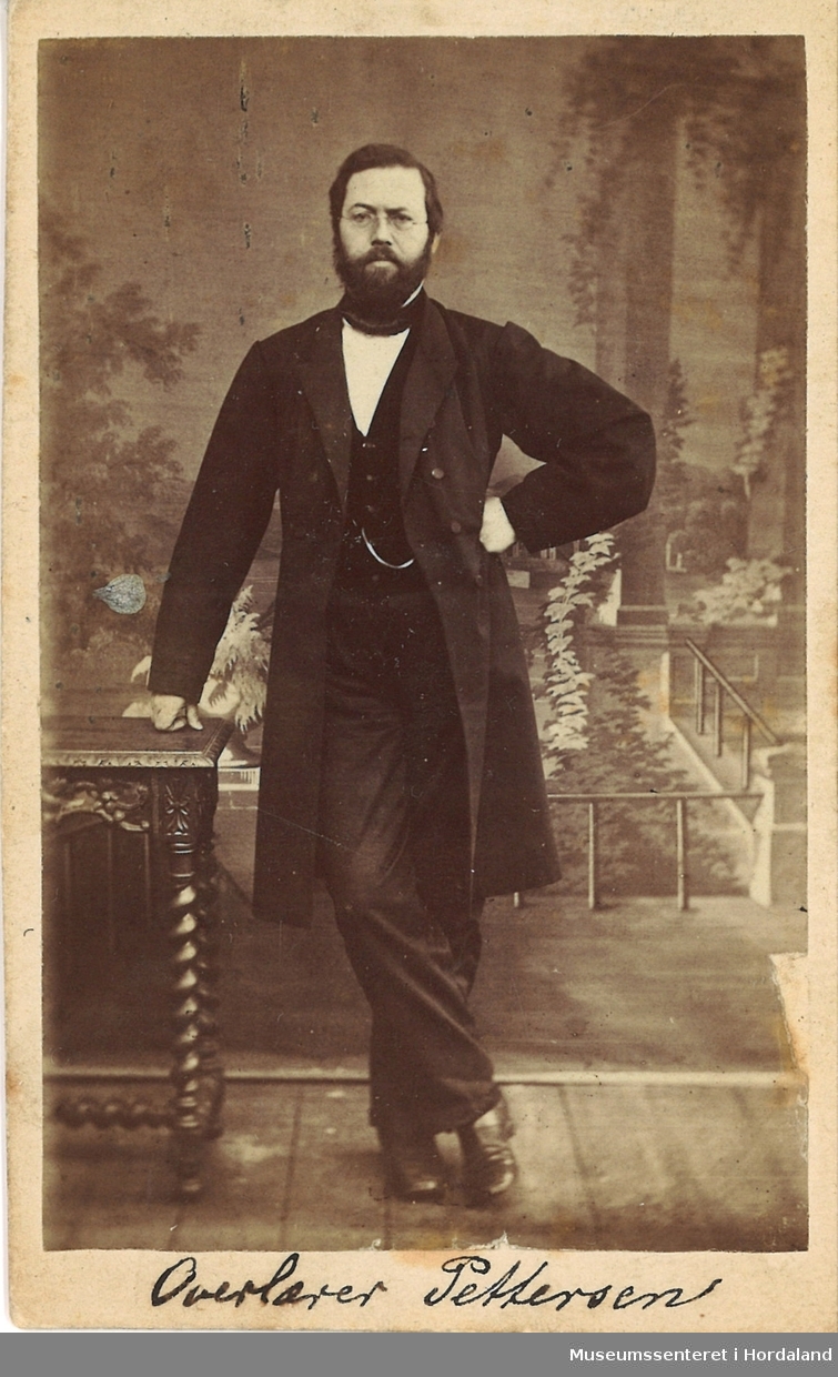 atelierfotografi av mann med skjegg i mørk dress med lang jakke, som lener seg til eit bord med høgre handa