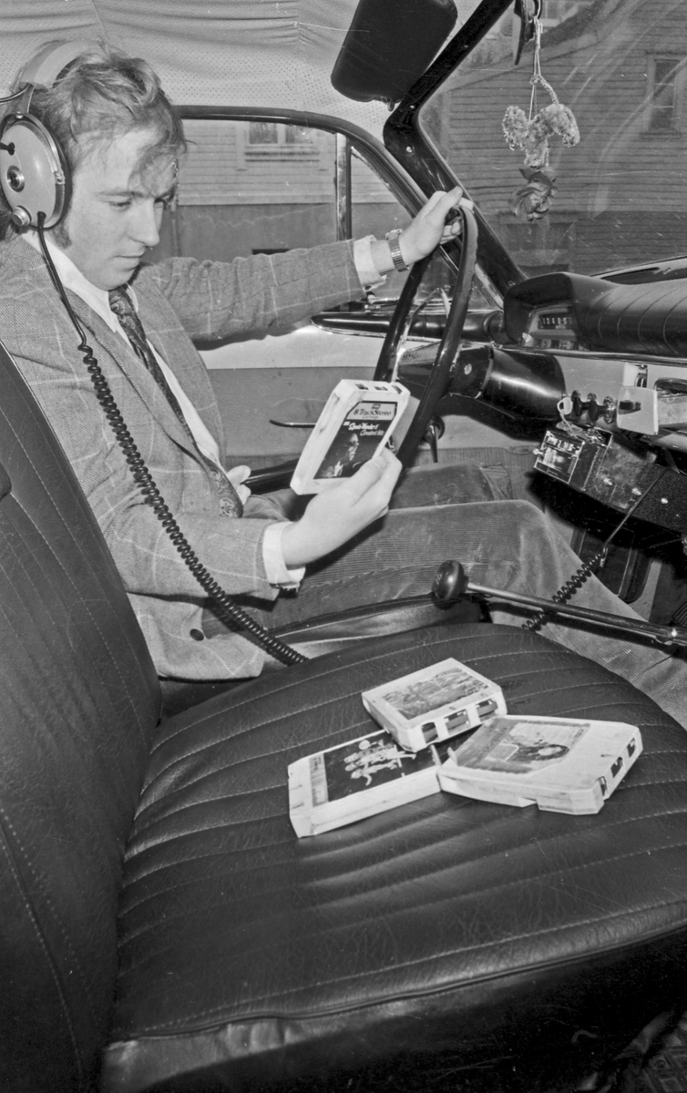 Mann med høreklokker. Testing av stereo i bil. Dyp konsentrasjon. Flere musikk-kassetter liggende på passasjersetet. Bilseteoanlegg av typen 8-spors. (Utdatert i dag i 2015.)