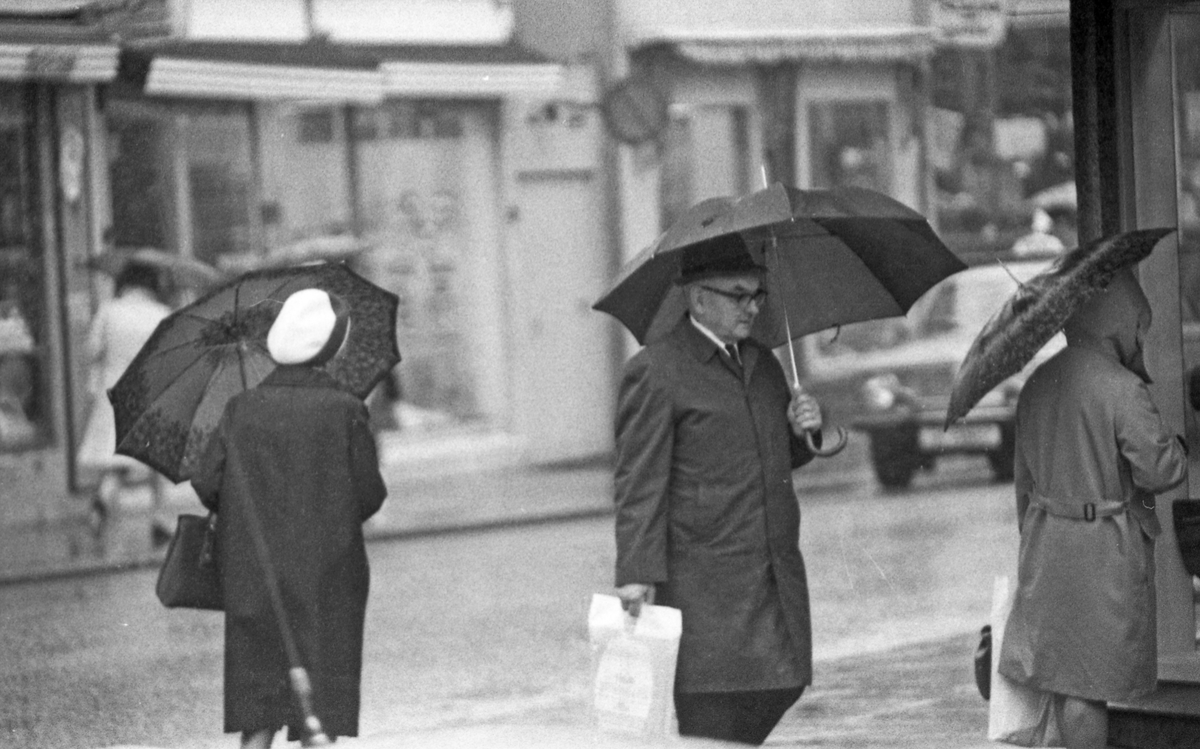Regnværsbilder. En travel bygate med paraplybærende innbyggere som haster avsted i regnværet.