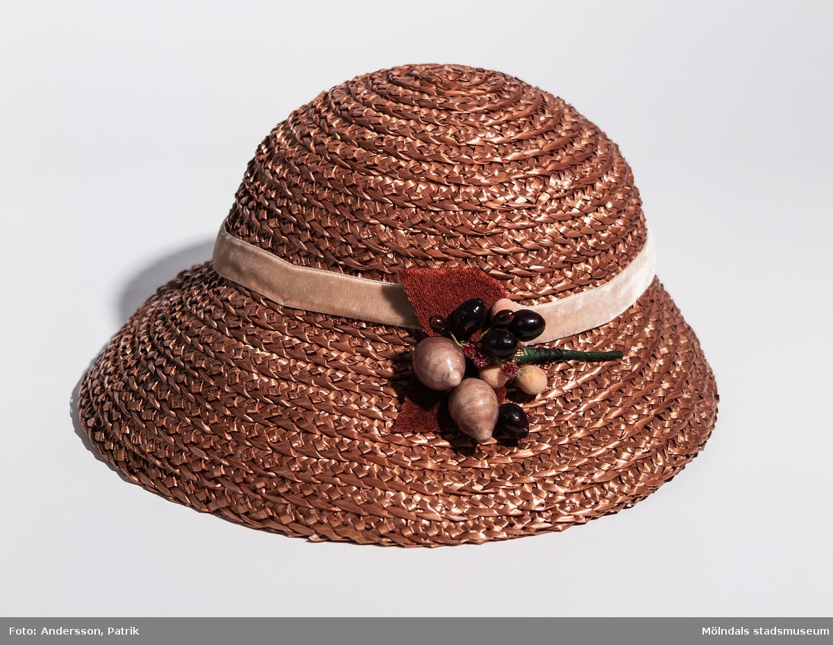 Damhatt inköpt i Eskilstuna, troligtvis på 1930-talet.
Hatten är ljusbrun och har ett ljusbrunt band runt kullen. Den har papier-maché dekoration fastsydd på vänster sida av hatten.
I hatten finns en pappersetikett fastsydd där siffrorna  
"4551 4229 696" står skrivna med blyerts.

Enligt uppgift från givaren är hatten inköpt på 1920-talet, men eftersom butiken öppnade 1934 är hatten troligen inköpt på 30-talet.